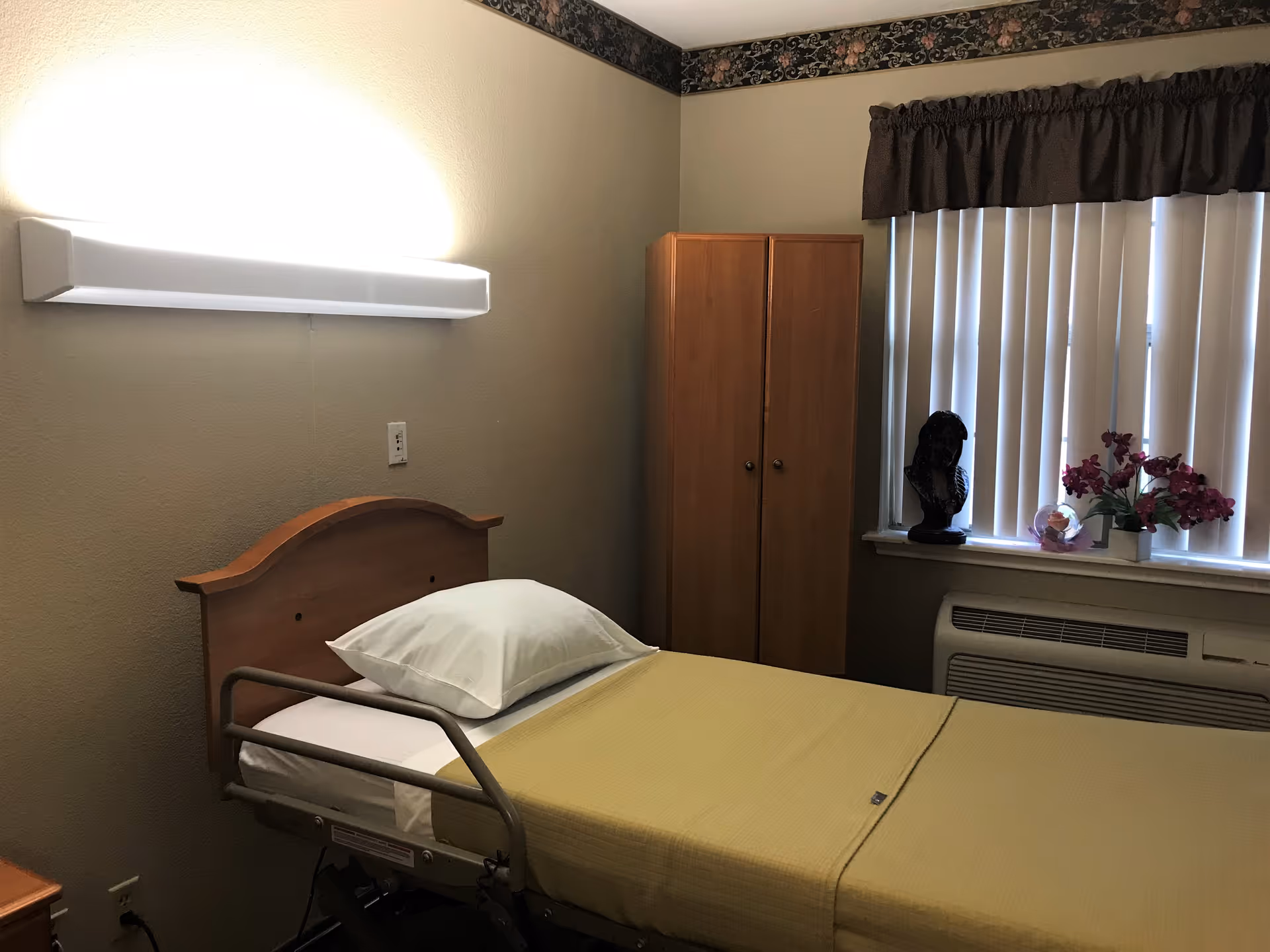 A simple senior care bedroom with a single hospital-style bed, wooden headboard, wardrobe and a window with vertical blinds and flowers.