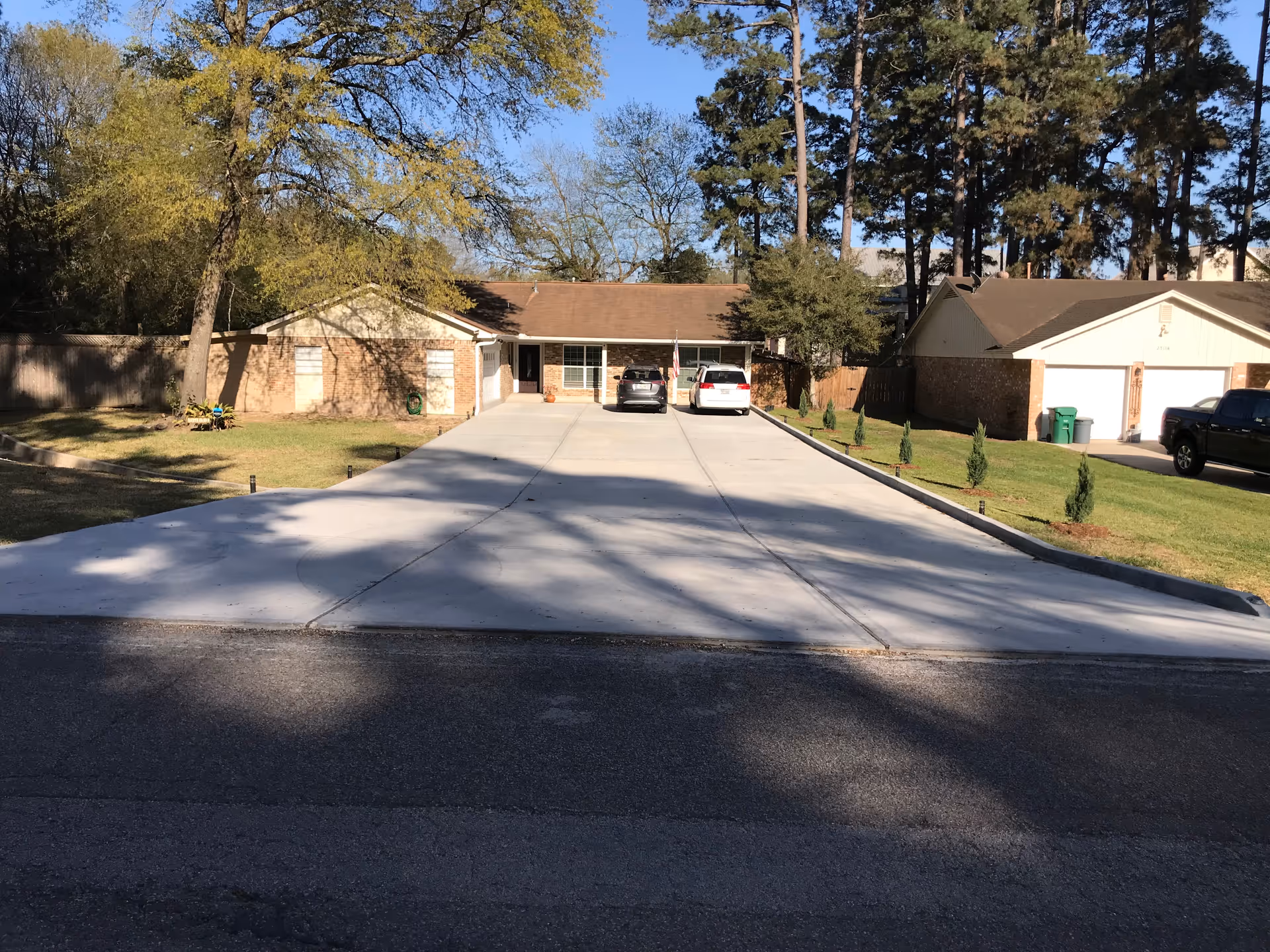 Front exterior view of a single-story brick building with a wide concrete driveway leading up to it. Two cars are parked in front of the building. There are trees and grass on either side of the driveway, and neighboring houses are visible.