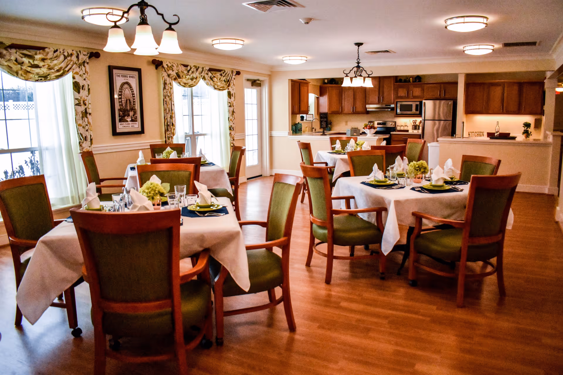 A dining room in a senior living facility with several tables covered in white tablecloths, each set with plates, glasses, napkins, and green floral centerpieces. The room has wooden floors, green cushioned wooden chairs, large windows with floral curtains, and a kitchen area in the background with wooden cabinets and stainless steel appliances.