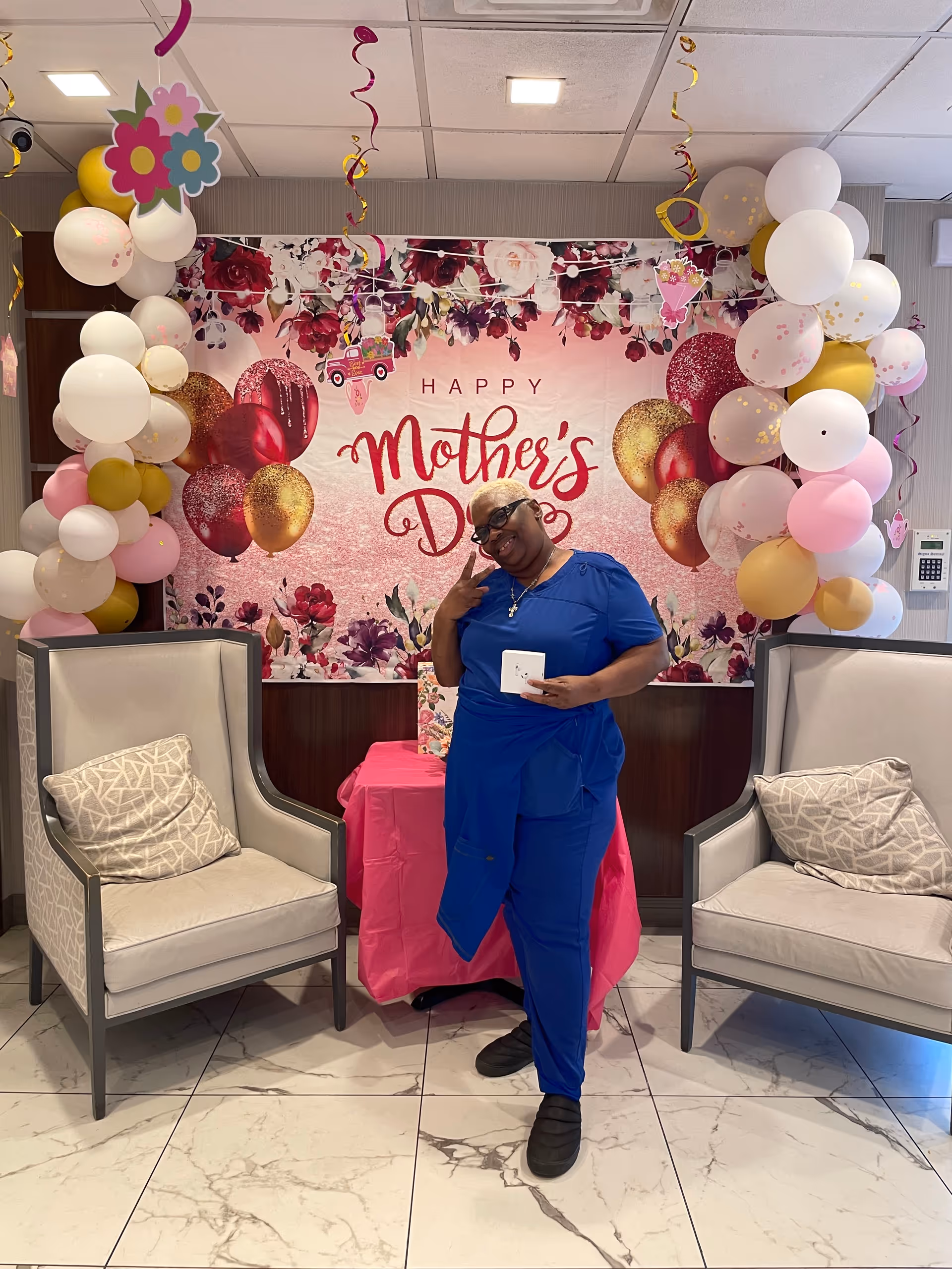 A woman dressed in blue scrubs poses smiling in front of a decorated backdrop that reads 'Happy Mother's Day'. The backdrop features floral and balloon designs in pink, red, and gold. There are balloon arches on either side of the backdrop with white, pink, and gold balloons. Two beige armchairs with patterned cushions are positioned on either side of the woman. The floor is tiled with a marble pattern.