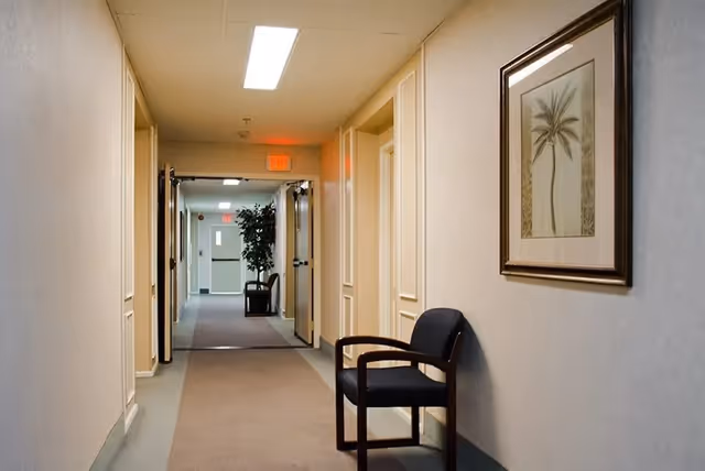 A well-lit hallway in a senior living facility with beige walls and carpeted floor. There is a black chair with wooden armrests placed against the right wall beneath a framed picture of a palm tree. Double doors are open at the end of the hallway, revealing another corridor with a potted plant and exit signs visible.