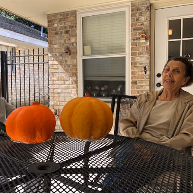 An elderly woman sitting at a black metal outdoor table with two pumpkins on it, under a covered patio attached to a brick building with a window and a door in the background.