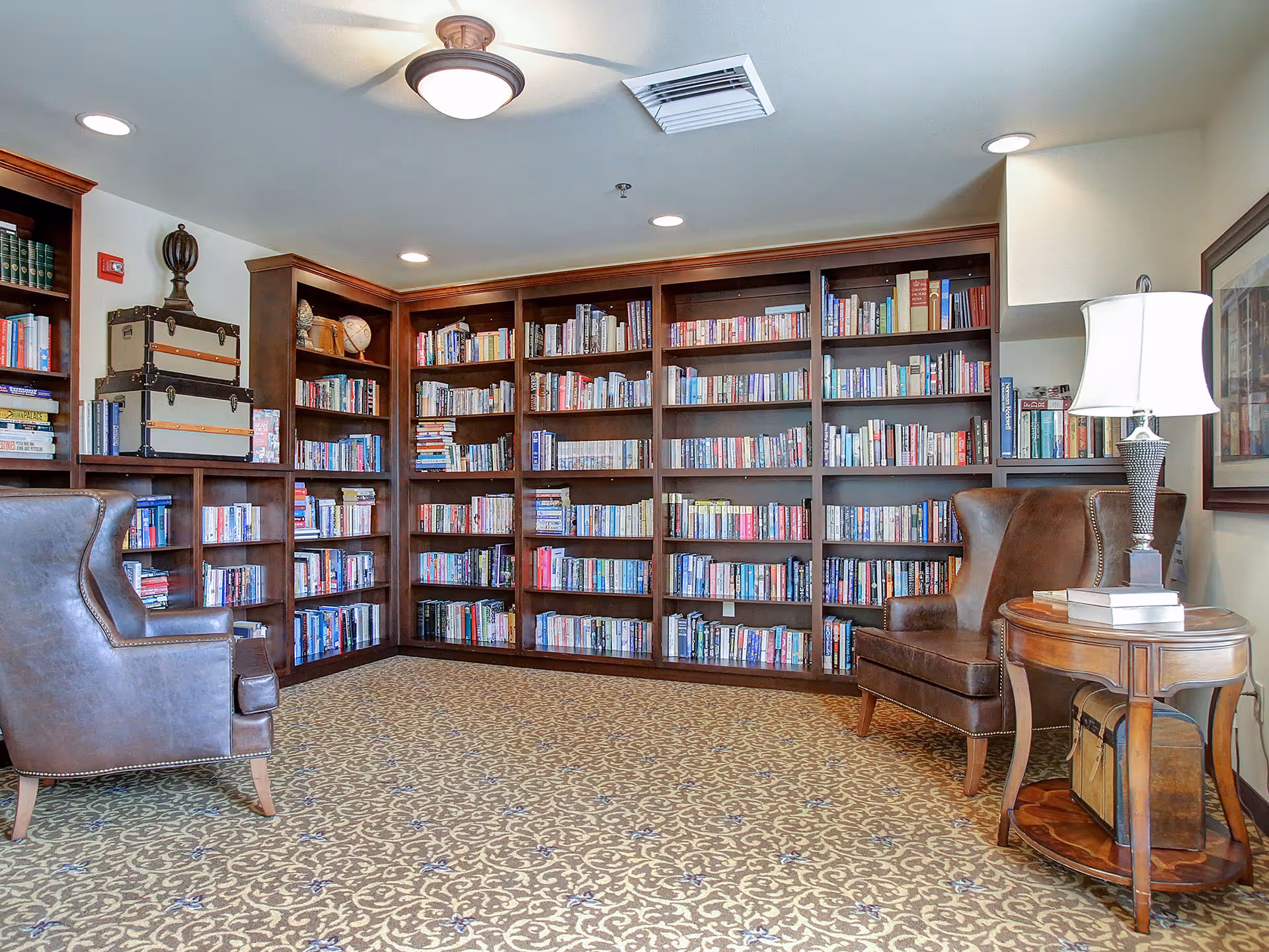 A cozy library room with wooden bookshelves filled with books along the walls. Two brown leather armchairs are positioned facing each other with a wooden side table between them, which holds a lamp and some books. The room has a patterned carpet and recessed ceiling lights.