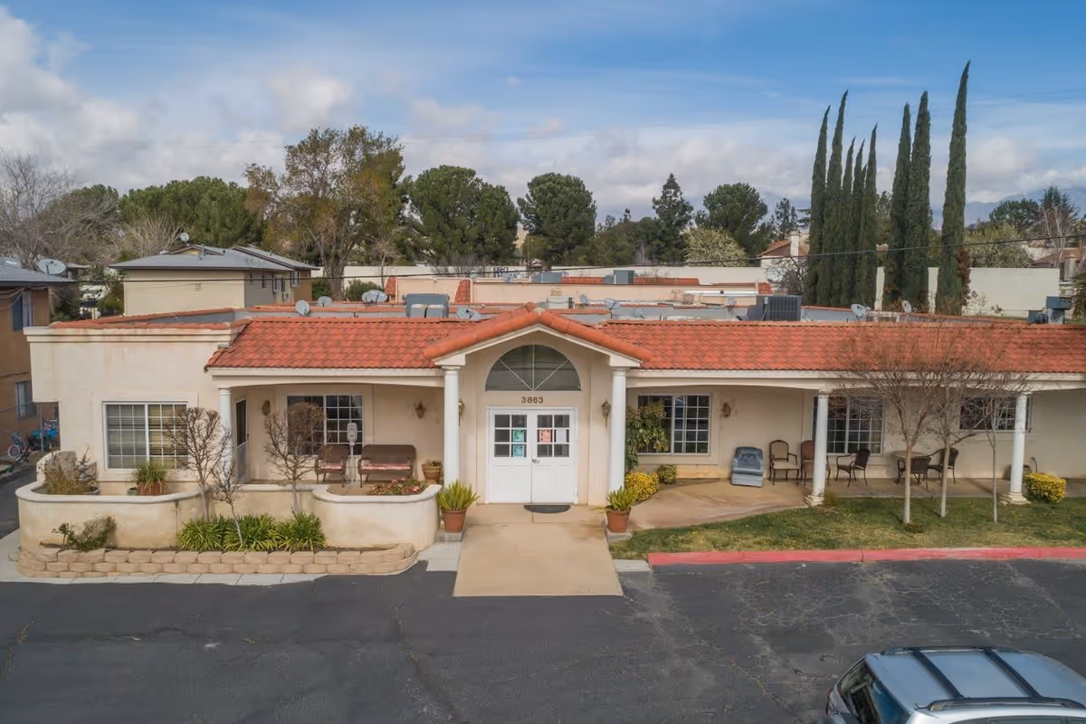 Front exterior view of a single-story building with a red tiled roof, white columns, and a central entrance door. There are several chairs and potted plants on the porch area, with trees and shrubs around the building. A parking lot is visible in the foreground under a partly cloudy sky.