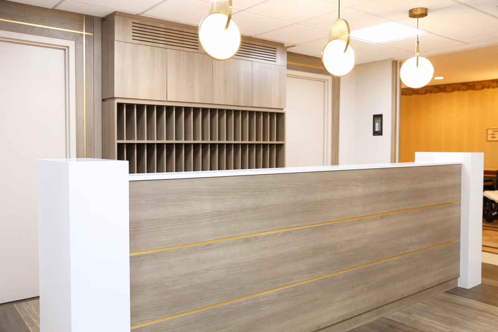 Reception desk in a senior living facility lobby with a wood-paneled counter, cubby mail slots behind it, and round pendant lights overhead.