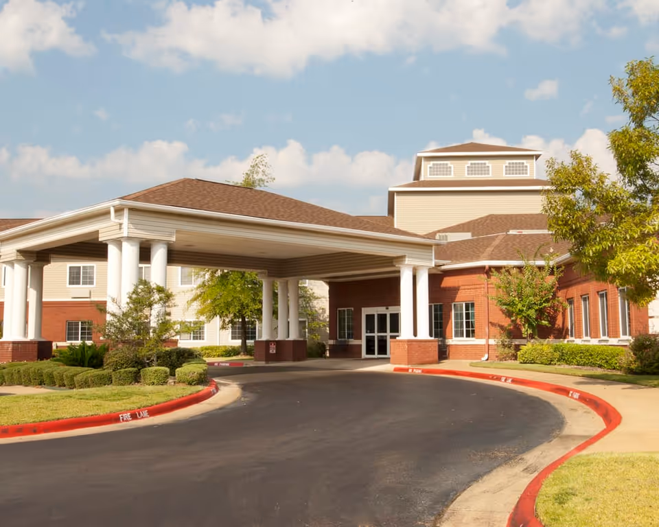 Exterior view of Garden Estates of Tyler, showing the main entrance with a covered driveway supported by white columns, surrounded by well-maintained landscaping and trees under a partly cloudy sky.