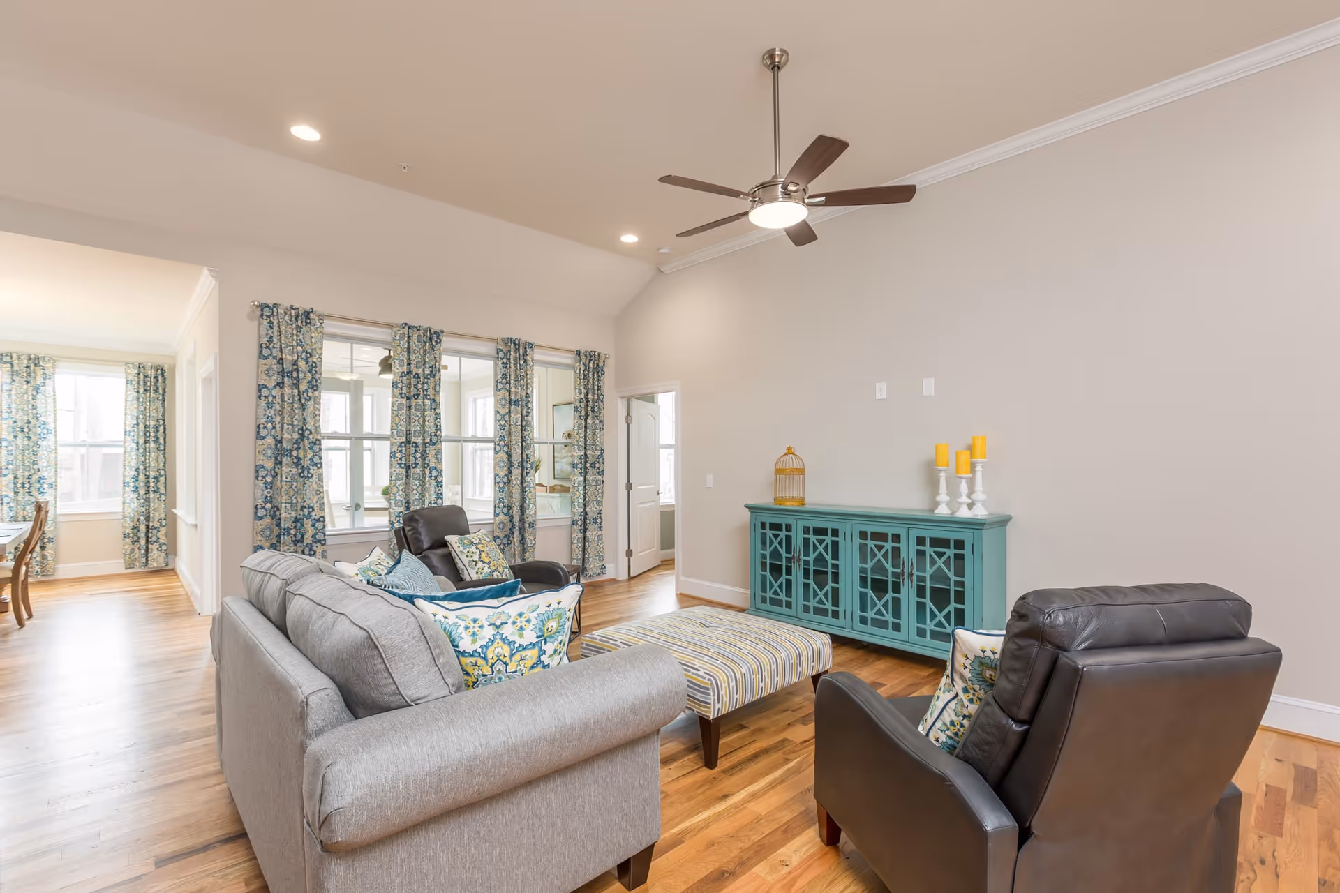A bright living room with hardwood floors, featuring a gray sofa with patterned pillows, two black leather armchairs, and a striped ottoman. A teal cabinet with decorative items, including a birdcage and candles, is against the wall. Blue and white patterned curtains hang on the windows, and a ceiling fan with lights is mounted on the ceiling.