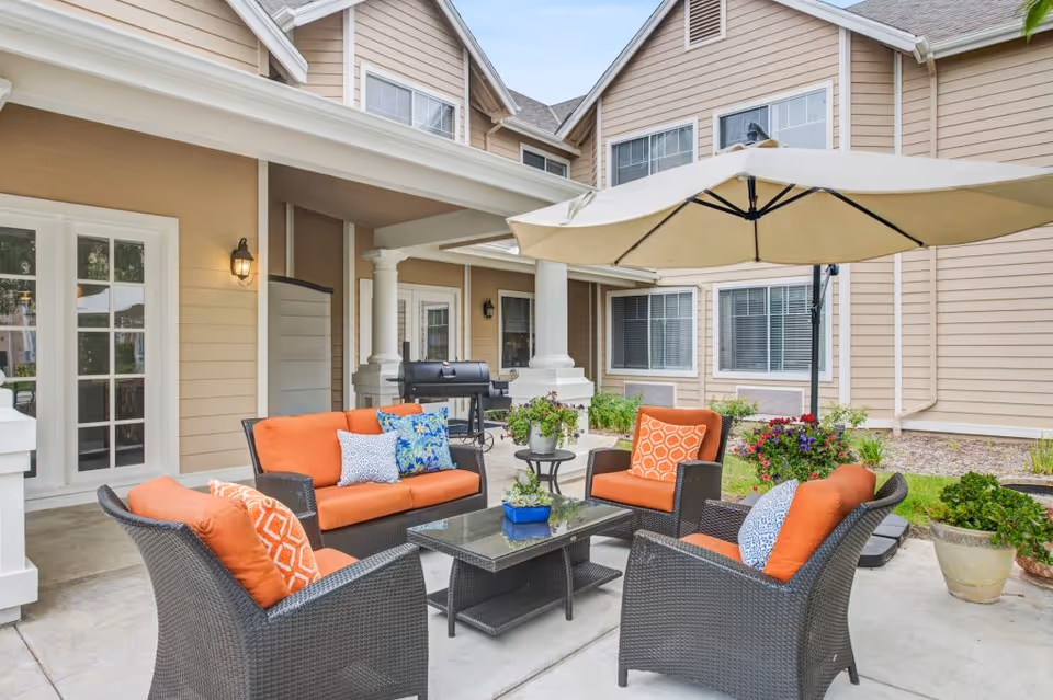 Outdoor patio area at Aegis Living Dana Point featuring a seating arrangement with four wicker chairs and a loveseat with orange cushions and decorative pillows around a glass coffee table. A large beige umbrella provides shade, and there are potted plants and flowers nearby. The background shows the beige exterior walls and windows of the building.
