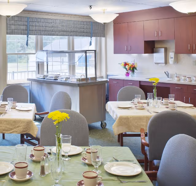 A bright dining room with several tables set with plates, cups, glasses, and silverware. Each table has a small vase with yellow flowers. In the background, there is a serving station with a sneeze guard and a countertop with cabinets, a paper towel dispenser, and a soap dispenser. Large windows let in natural light.
