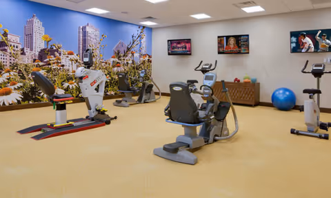 A senior living facility exercise room with several stationary exercise bikes and fitness equipment. The room has a large mural of a city skyline with flowers on one wall and three televisions mounted on another wall. The floor is carpeted and the room is well-lit with ceiling lights.