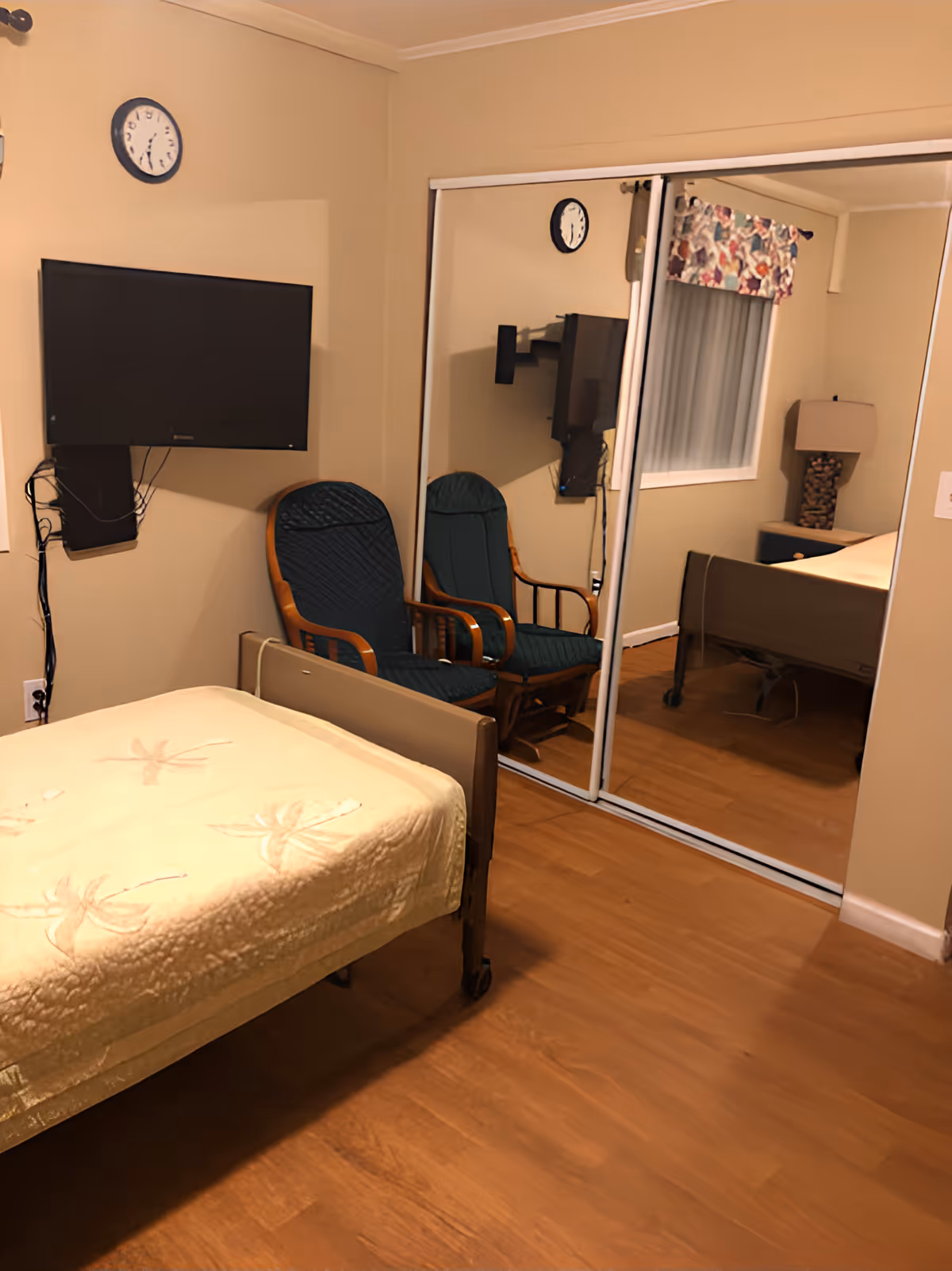 A bedroom in an assisted living home with a single bed covered with a light yellow blanket, a wall-mounted flat-screen TV, a clock above the TV, two wooden chairs with dark cushions, a mirrored sliding closet door reflecting part of the room, a window with a colorful valance, and a bedside table with a lamp.