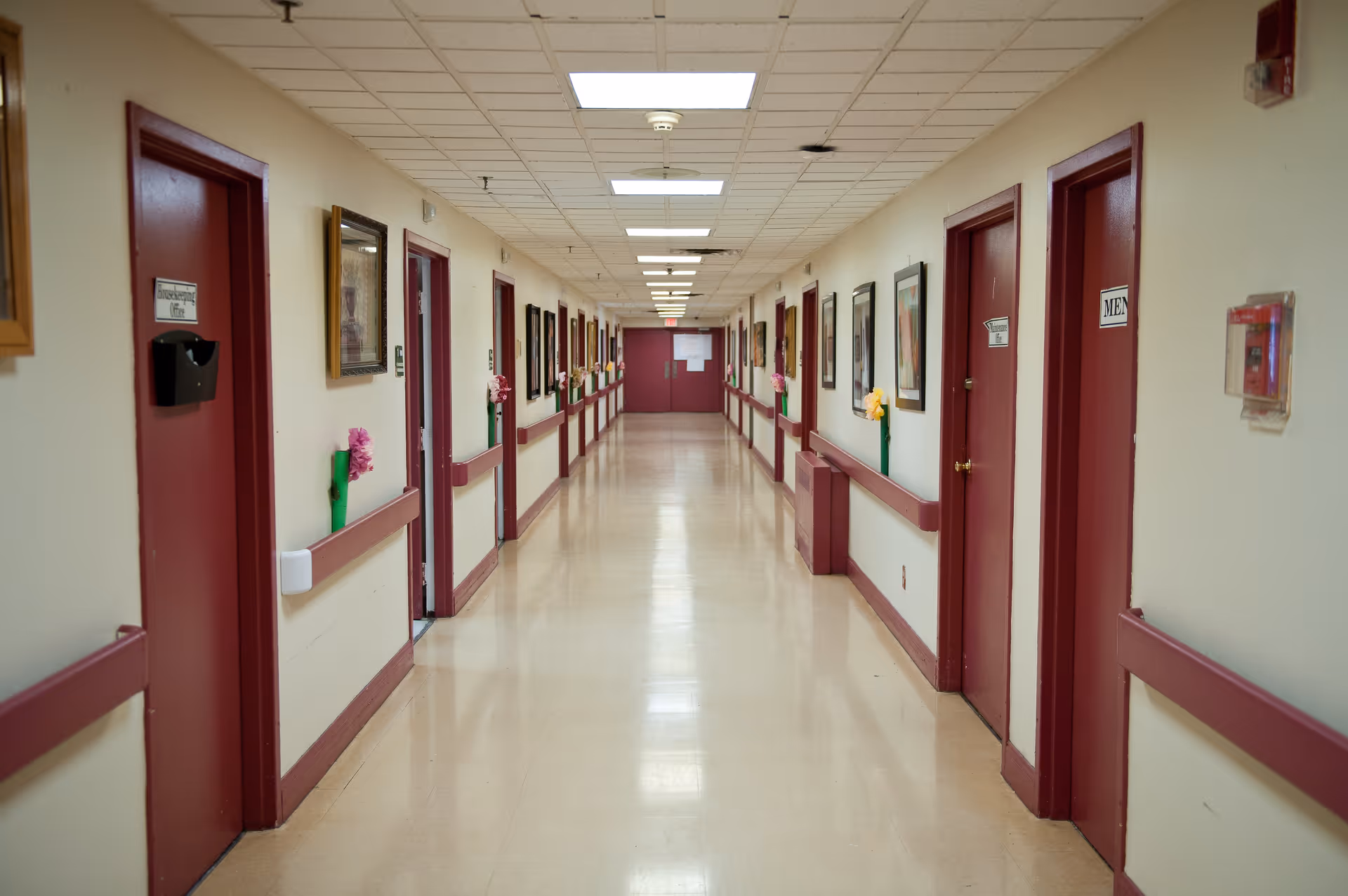 A long, clean hallway in a healthcare facility with maroon doors on both sides, beige walls, handrails, framed pictures, and ceiling lights.