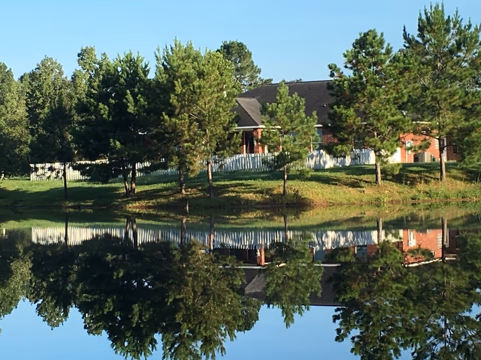 A serene outdoor scene featuring a row of pine trees and a brick building with a dark roof behind a white picket fence, all reflected clearly in a calm body of water in the foreground under a clear blue sky.