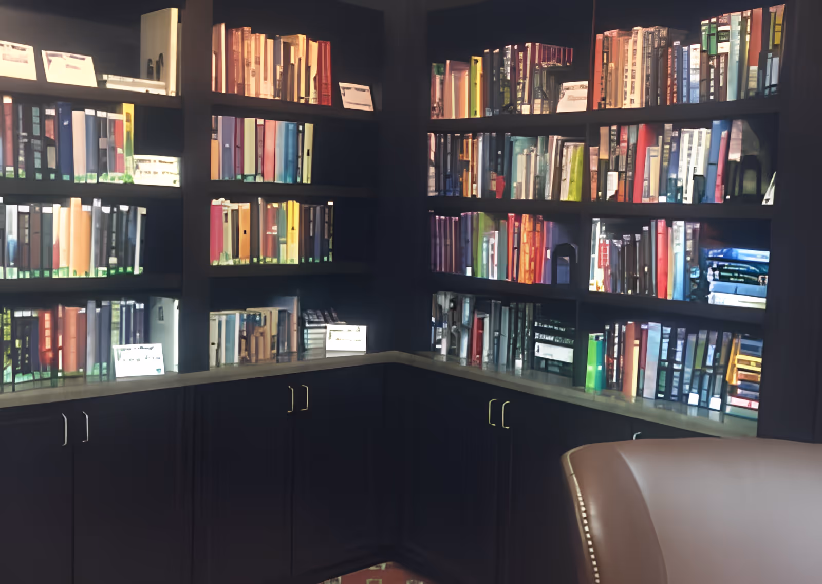 Corner bookshelf filled with numerous colorful binders and books, with dark cabinetry below and a partial view of a beige leather chair in the foreground.