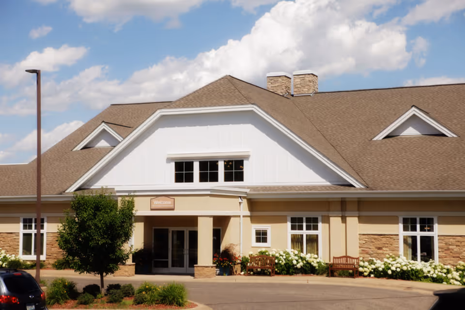 Front exterior of Ecumen North Branch showing the main entrance with benches, landscaping, and a pitched roof under a partly cloudy sky.