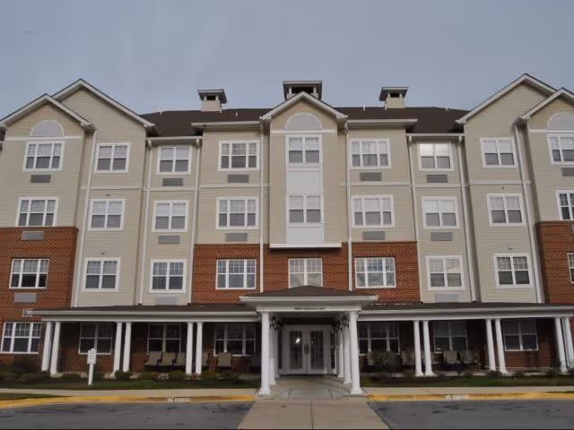 Front exterior view of a multi-story senior living facility building with beige siding and red brick accents, featuring multiple windows and a covered entrance with white columns.