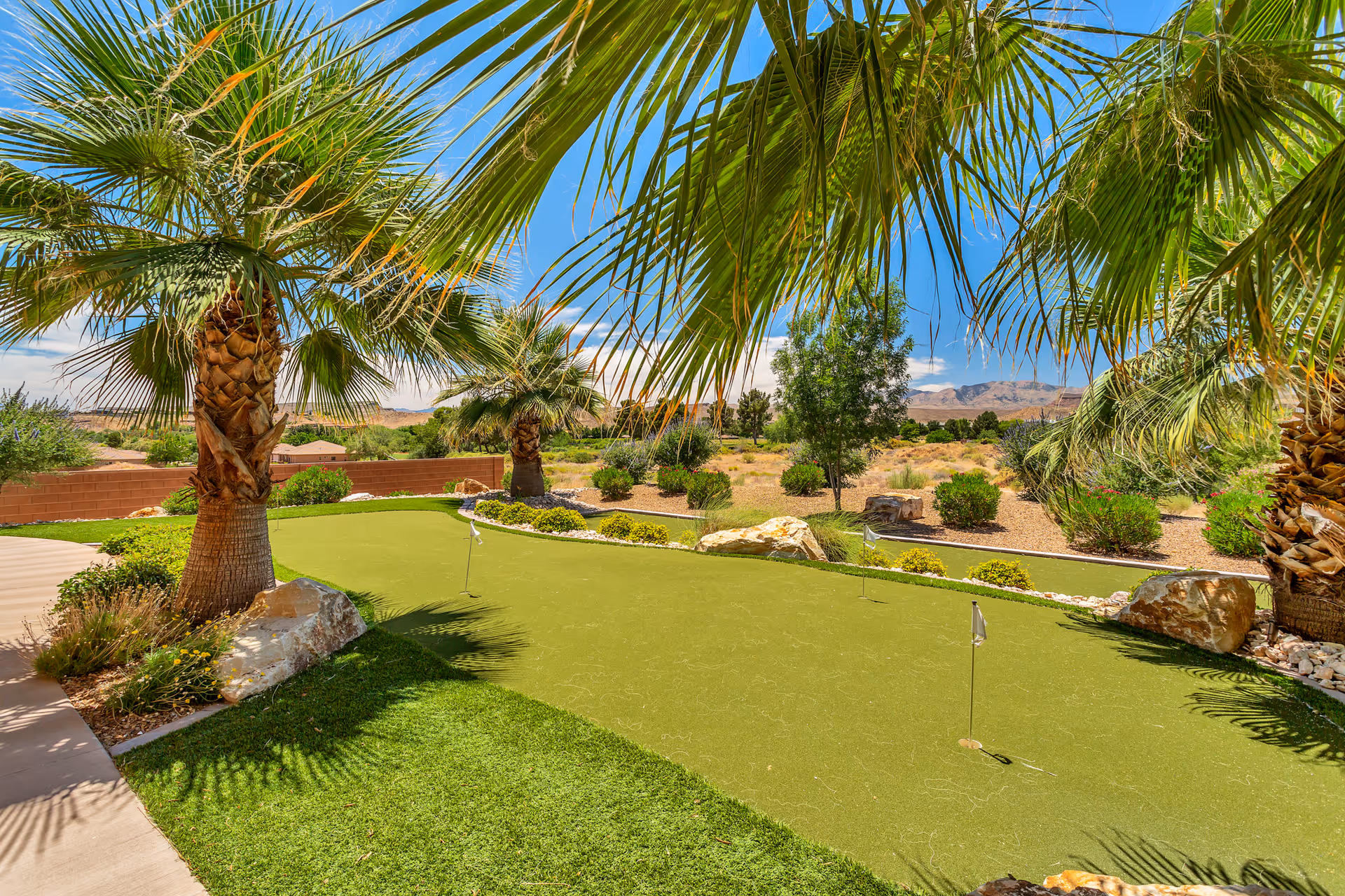 Outdoor putting green surrounded by palm trees and desert landscaping under a clear blue sky with mountains in the background.