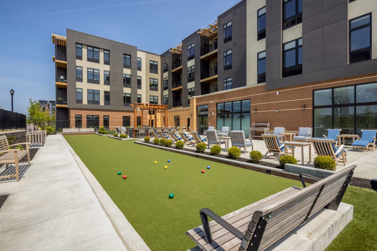 A modern multi-story building with an outdoor bocce lawn, benches and lounge chairs on a sunny day.