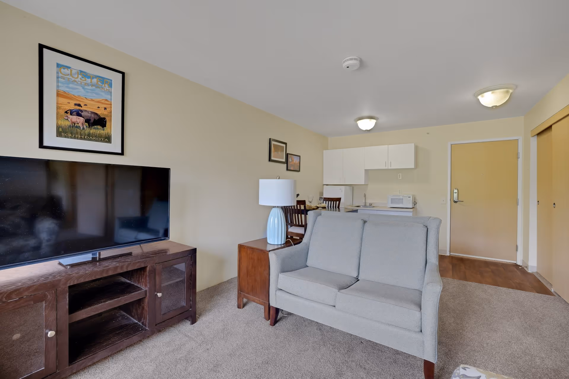Interior view of a senior living apartment featuring a gray loveseat, a wooden TV stand with a flat-screen television, a side table with a blue lamp, a small dining table with chairs, and a kitchenette with white cabinets, a microwave, and a refrigerator. The room has beige walls, carpeted flooring, and a wooden door leading to the entrance.