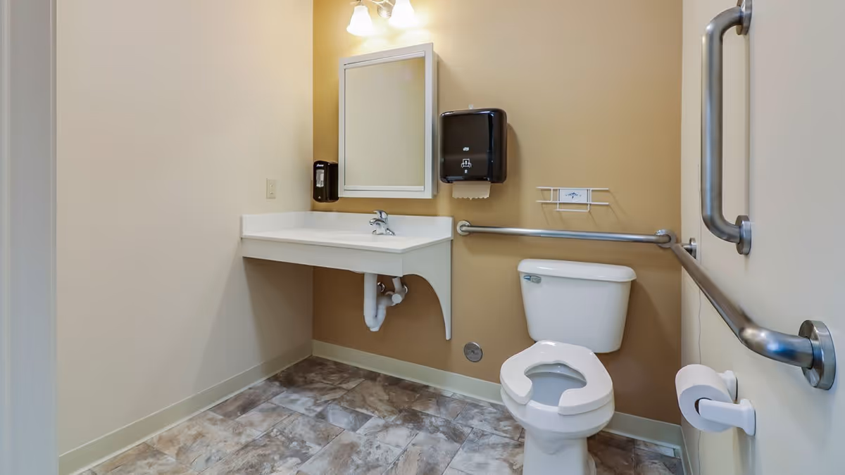 Accessible bathroom with beige walls and tiled floor featuring a white toilet with safety grab bars on the walls, a wall-mounted white sink with a mirror above it, a soap dispenser, and a paper towel dispenser.