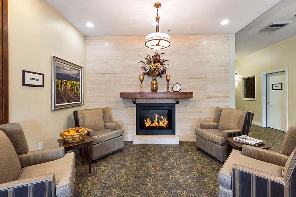 A cozy lounge area with four beige armchairs arranged around a small wooden table with a basket of fruit. A modern fireplace with a wooden mantel is centered on a tiled wall, decorated with a floral arrangement, two gold candle holders, and a small clock. The room has carpeted flooring and soft lighting from a ceiling fixture. Signs on the walls indicate the lounge and a salon door in the hallway.