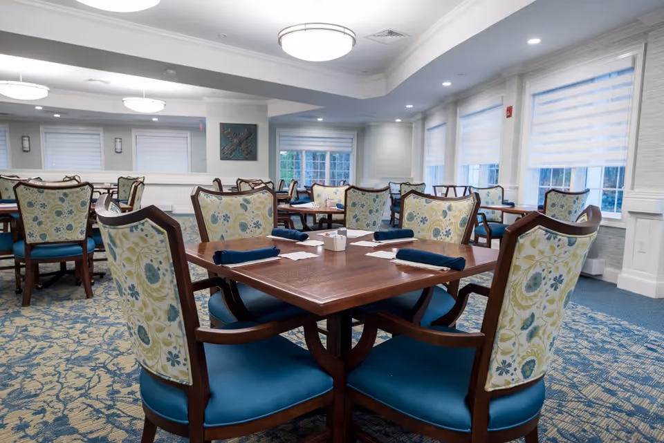 A dining room with several wooden tables and chairs featuring floral patterned upholstery and blue cushions. Each table is set with blue napkins and white napkin holders. The room has large windows with white blinds, light-colored walls, and ceiling lights providing bright illumination.