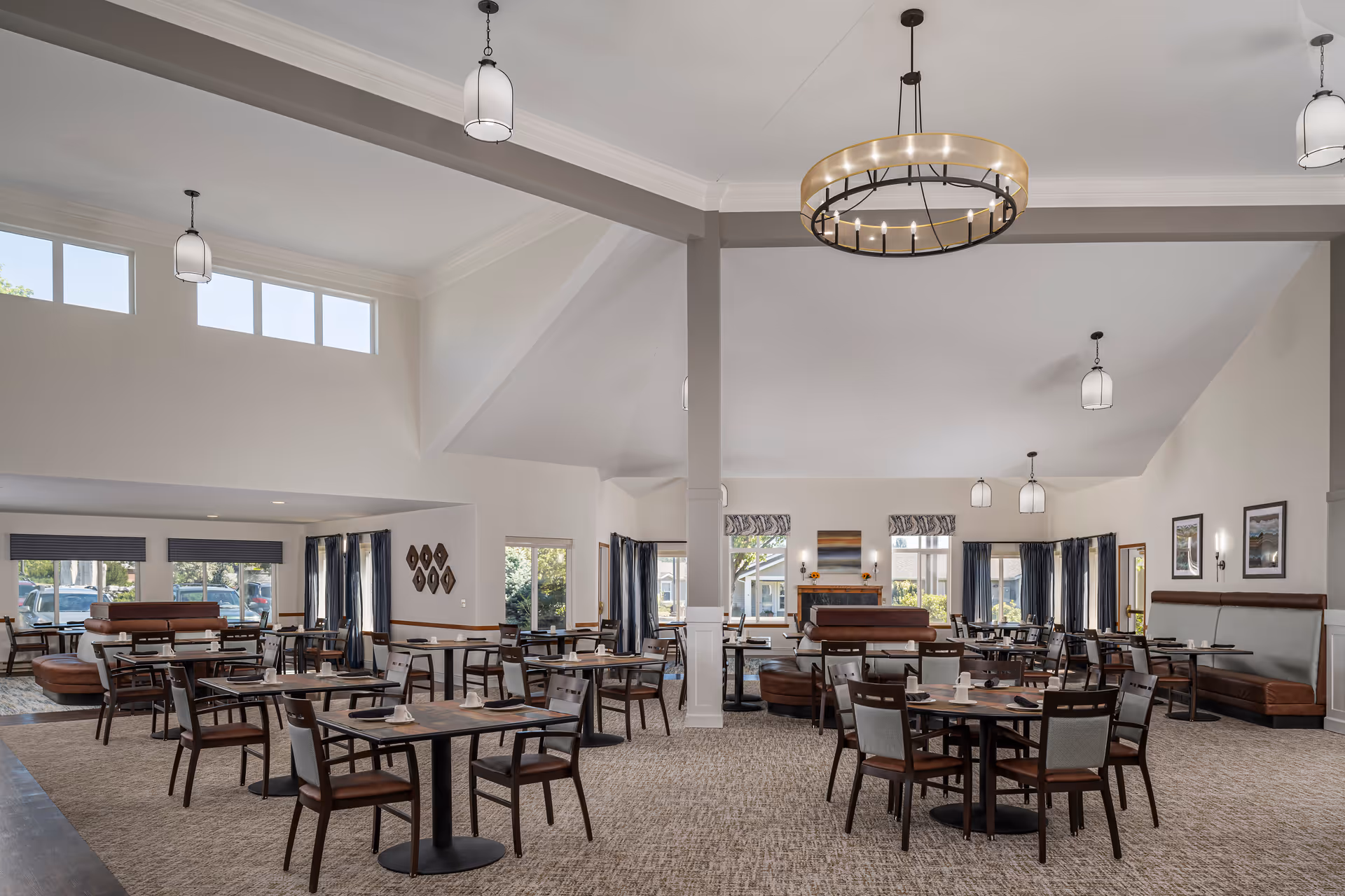 Spacious dining room in a senior living facility with multiple tables and chairs arranged neatly. The room features high ceilings with modern pendant lighting, large windows allowing natural light, and a cozy fireplace at the far end. The decor includes neutral tones with dark wood furniture and patterned curtains.