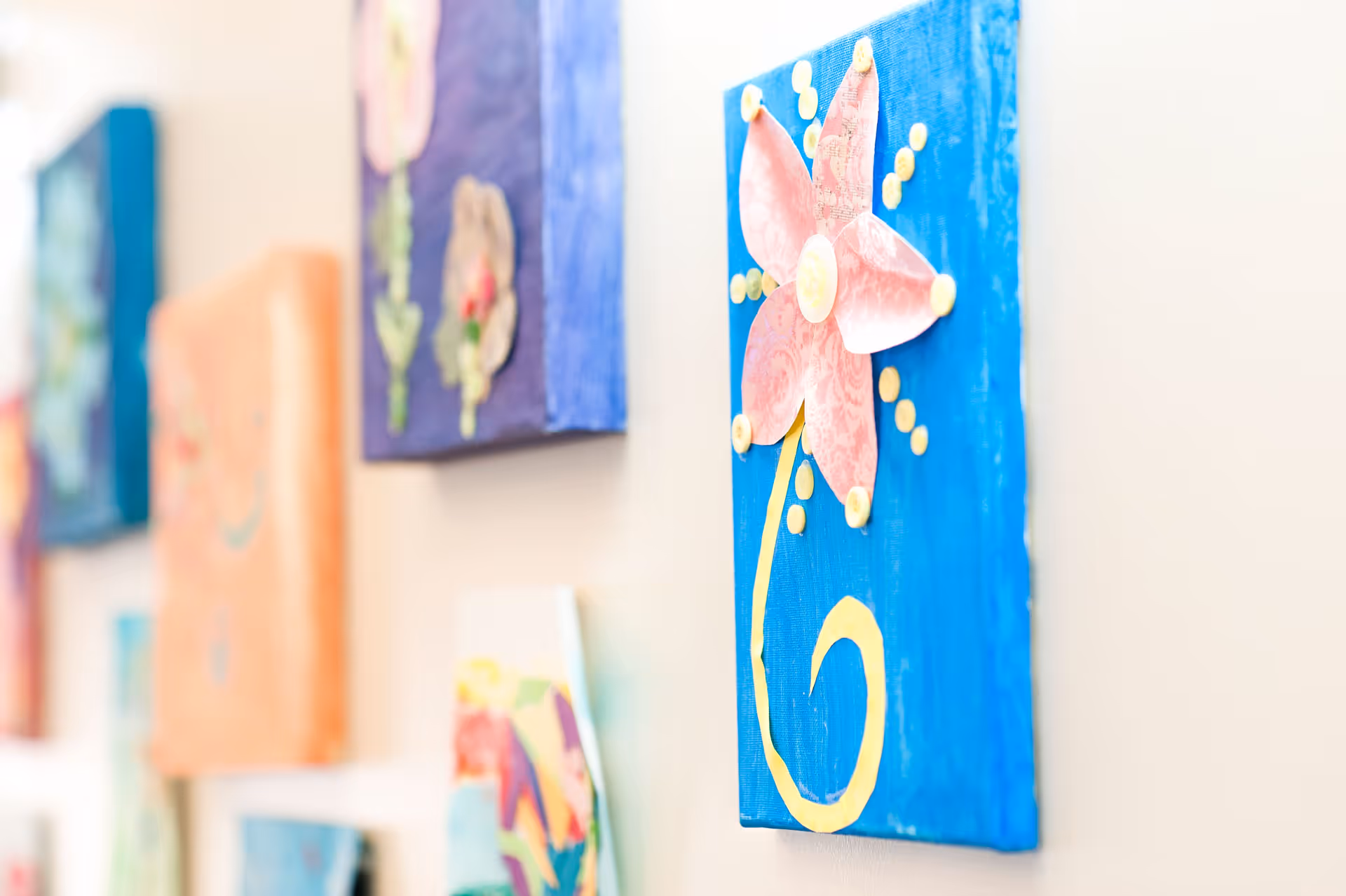 Close-up view of colorful, handmade flower artwork displayed on a wall, with a prominent blue canvas featuring a pink flower with yellow accents in focus.