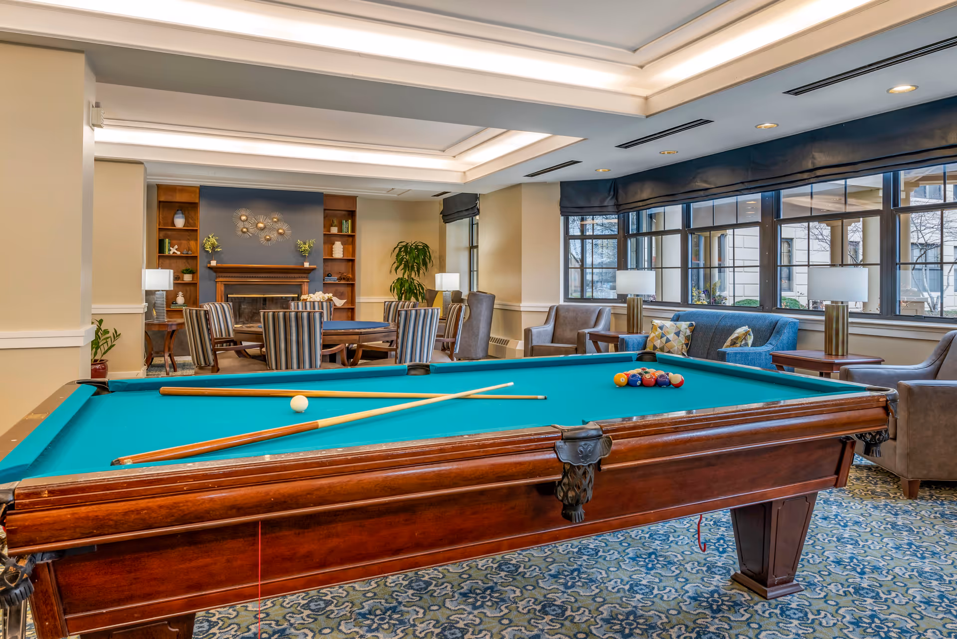 A well-lit recreational room featuring a pool table with cues and balls set up, surrounded by comfortable seating including armchairs and a sofa. In the background, there is a fireplace with built-in wooden shelves on either side, decorated with plants and ornaments. Large windows allow natural light to fill the room.