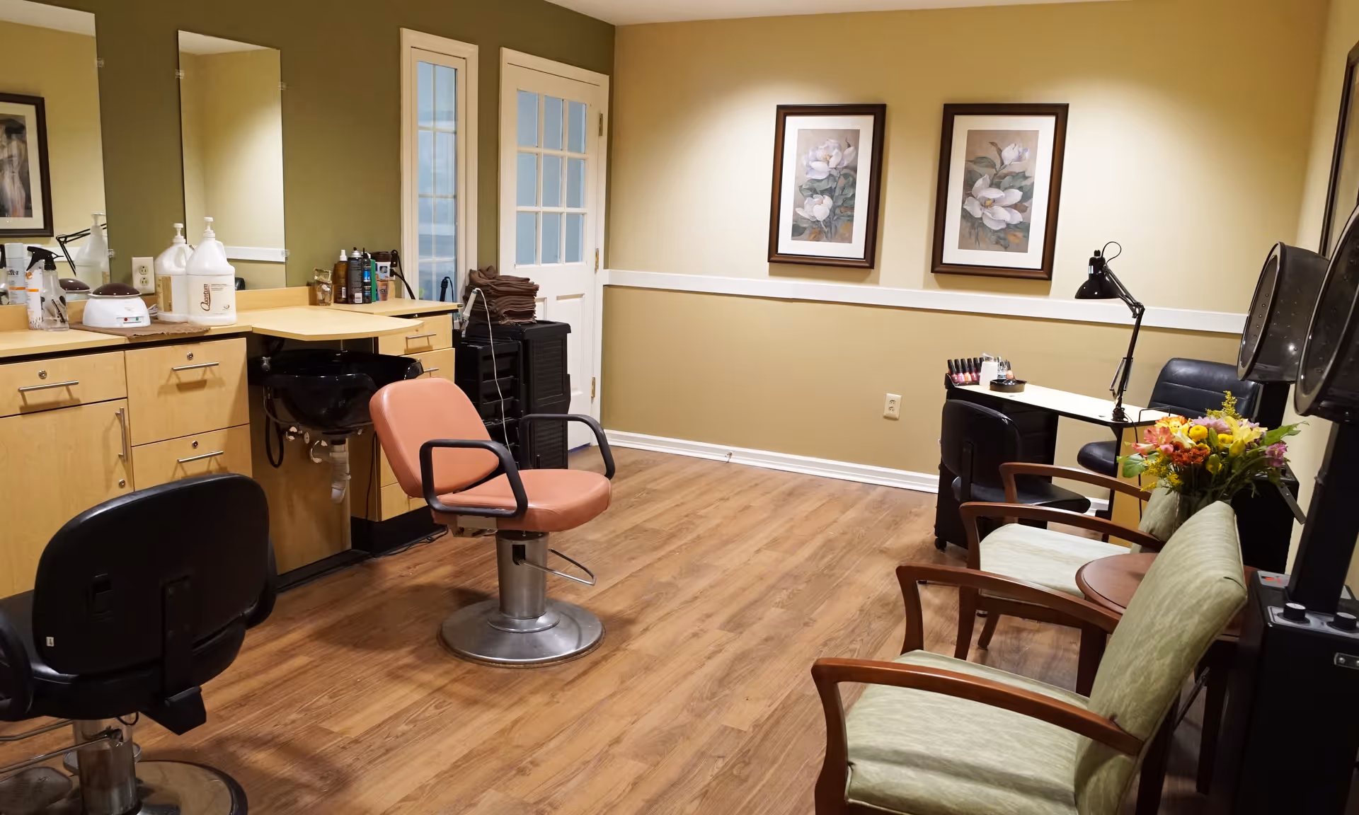 Small salon room with styling chairs, mirrors, cabinetry, and a seating area with framed floral prints.