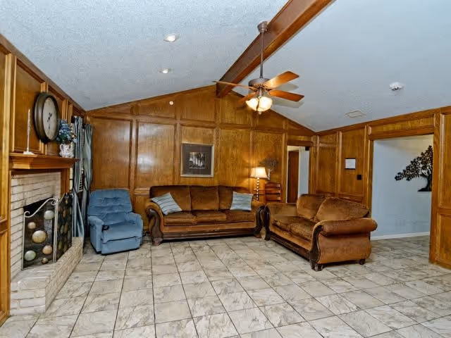 A cozy living room with wood-paneled walls, a tiled floor, a ceiling fan with lights, a fireplace with a clock on the mantel, a blue recliner chair, a brown sofa, and a brown loveseat. There is a lamp on a side table between the sofas and a framed picture on the wall.