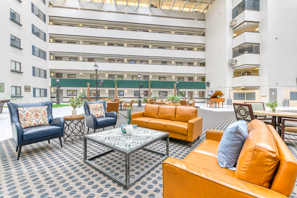 Spacious indoor common area with modern seating including two brown leather sofas, two blue armchairs with patterned pillows, and a glass coffee table. The area is carpeted with a patterned design and surrounded by multiple floors of the building with balconies overlooking the space. There are plants, decorative lamp posts, and a piano in the background under a large skylight ceiling.