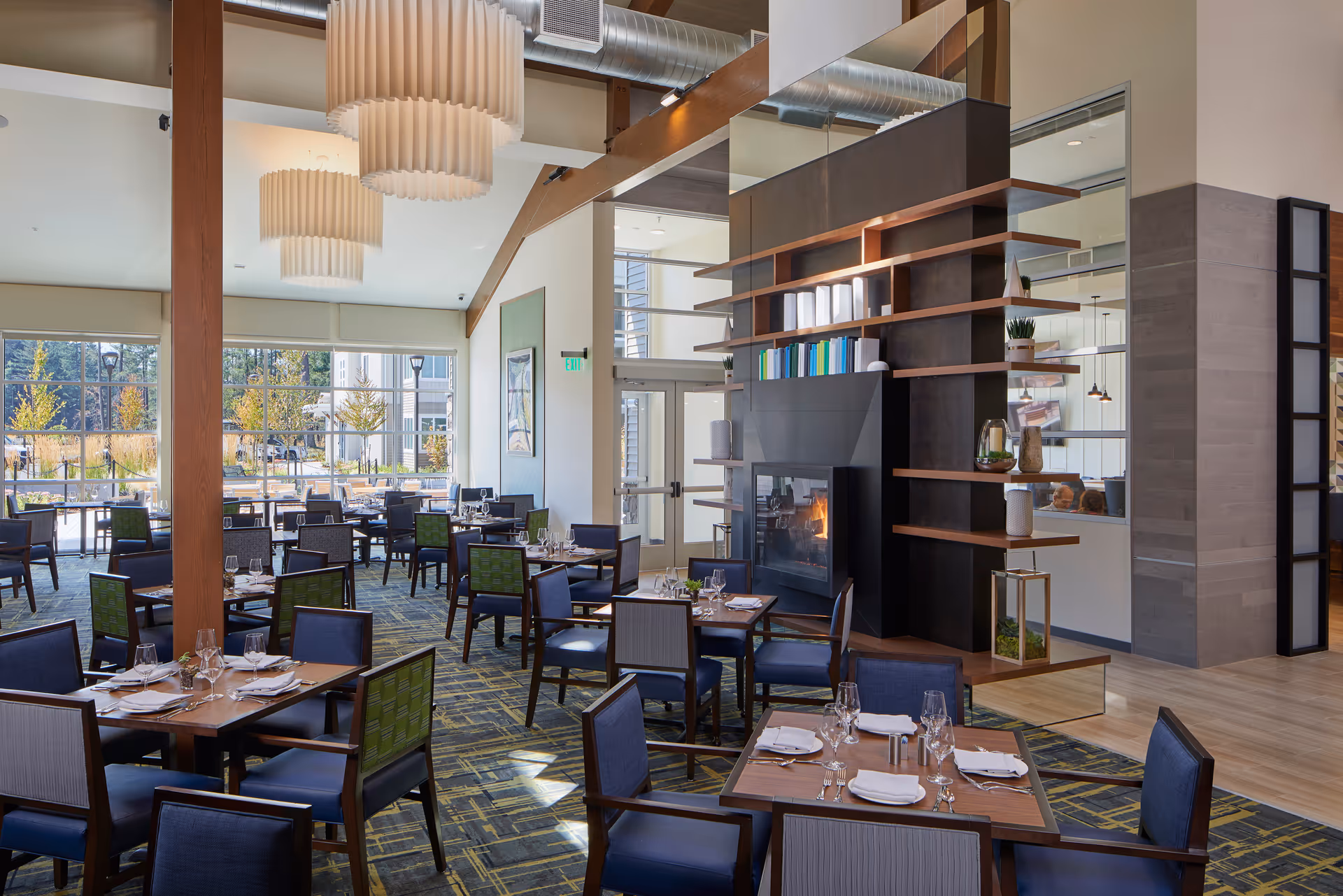 A modern dining room with multiple tables set with glasses, napkins, and cutlery. The room features large windows allowing natural light to fill the space, contemporary hanging light fixtures, a fireplace with decorative shelving, and a patterned carpet. Outside the windows, trees and outdoor seating are visible.