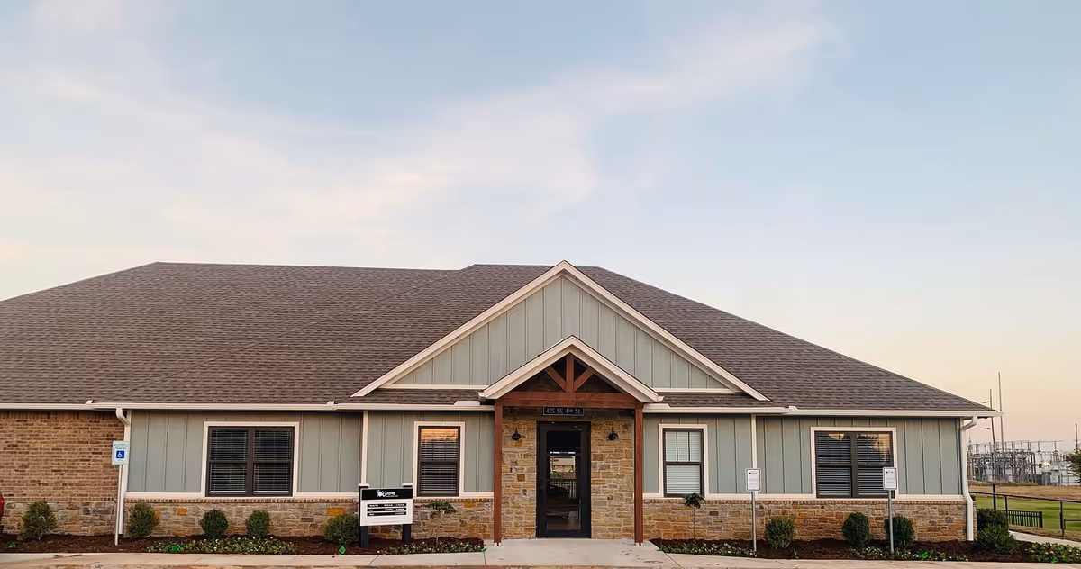 Exterior view of a single-story building with a gabled roof, stone and light green siding, and a central entrance with a small covered porch. There are windows on either side of the entrance and a landscaped area with small bushes in front. The sky is clear with soft evening light.