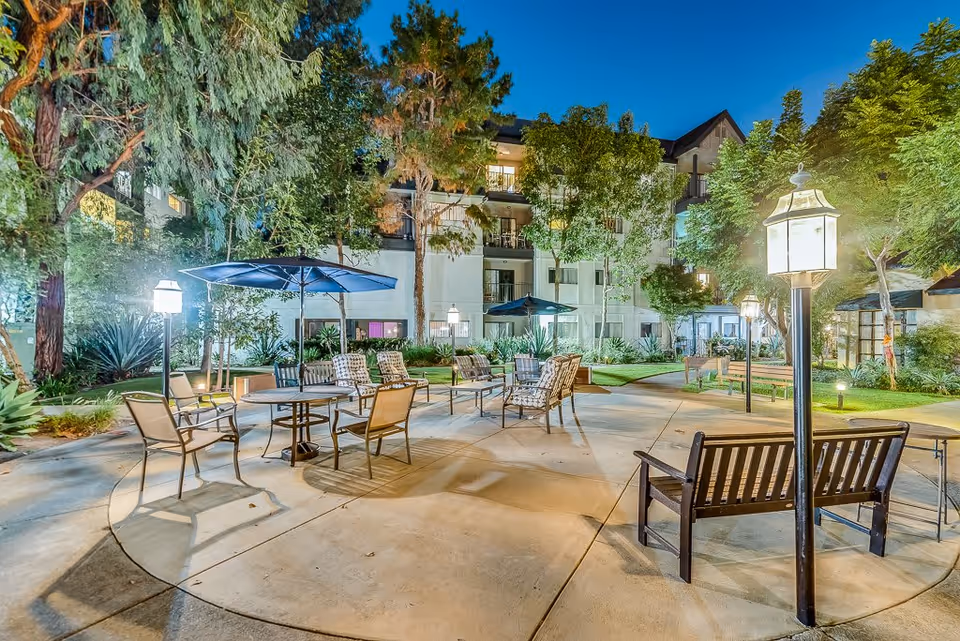 Outdoor patio area at night with multiple seating arrangements including chairs, tables with umbrellas, and benches surrounded by trees and greenery. The building of Westminster Terrace Senior Living is visible in the background with lit windows.