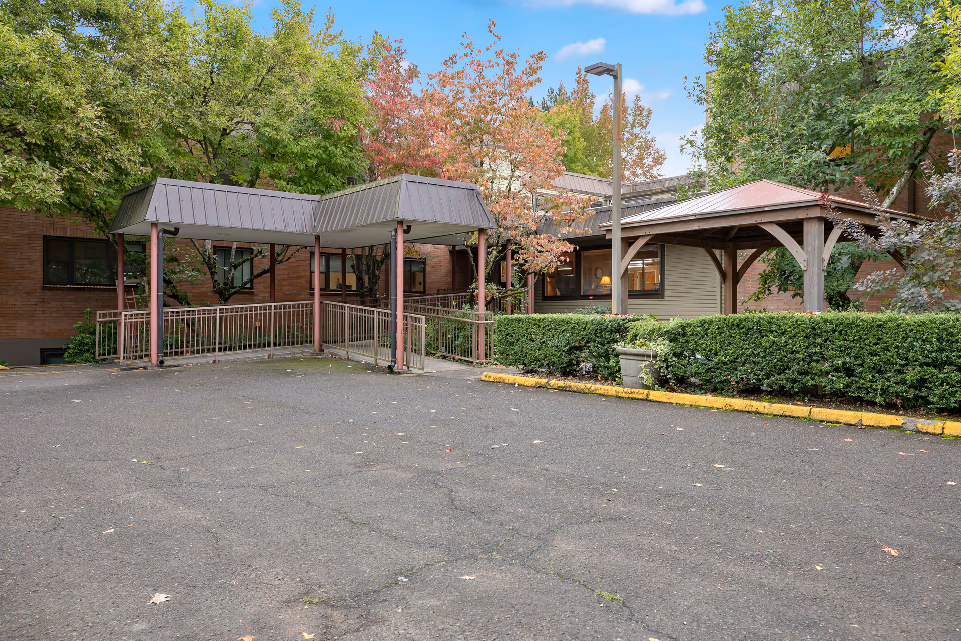 Outdoor view of a covered walkway and a wooden gazebo structure surrounded by trees with green and autumn-colored leaves, adjacent to a brick building with windows, and a paved area in the foreground.