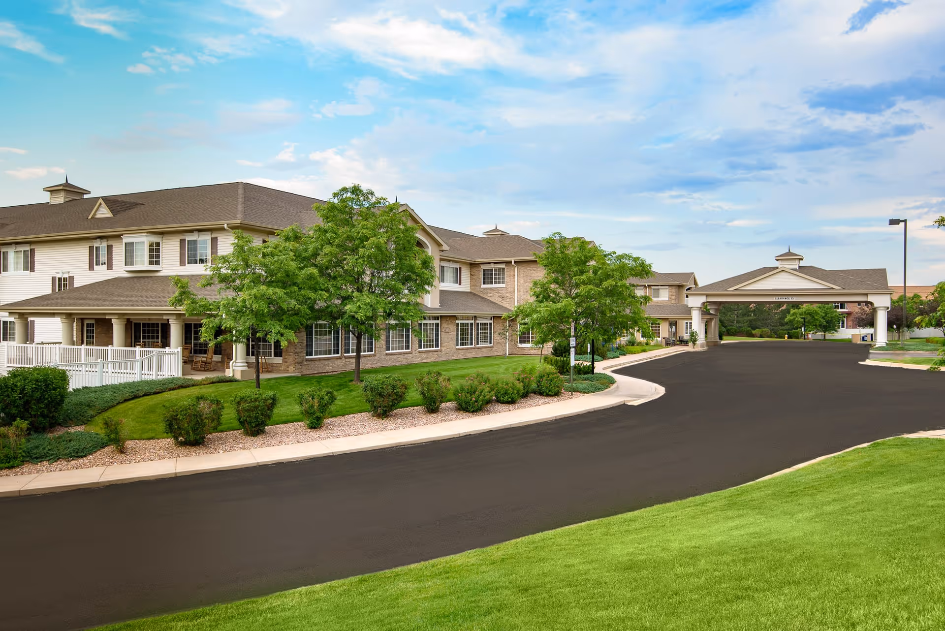Exterior view of a senior living facility named Inn at Garden Plaza showing a large two-story building with beige siding and brick accents, surrounded by green lawns, trees, and shrubs under a partly cloudy blue sky. There is a paved driveway leading to a covered entrance with columns.