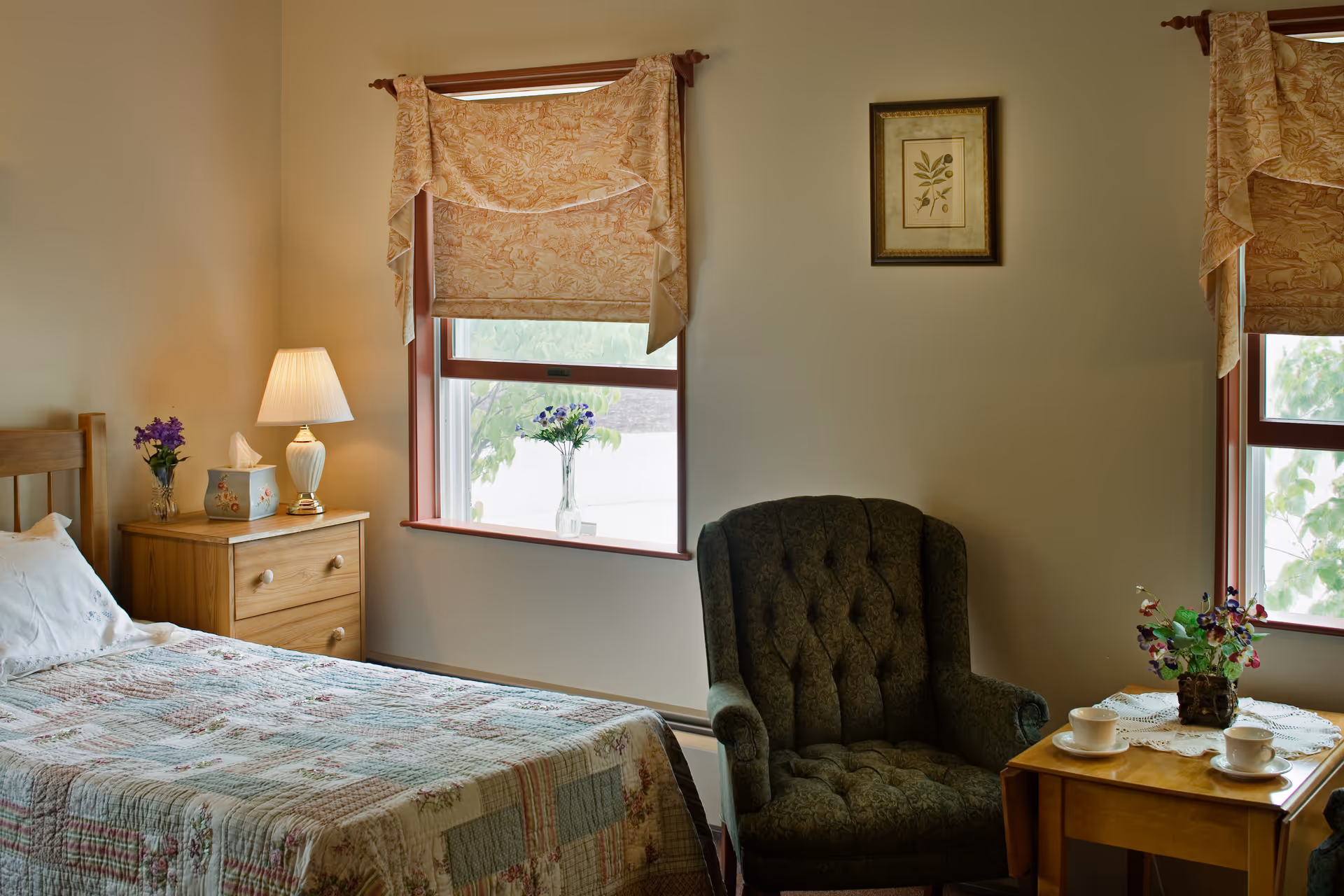 A cozy bedroom with a bed, wooden nightstand with a lamp and flowers, an upholstered armchair, and a side table by two windows.