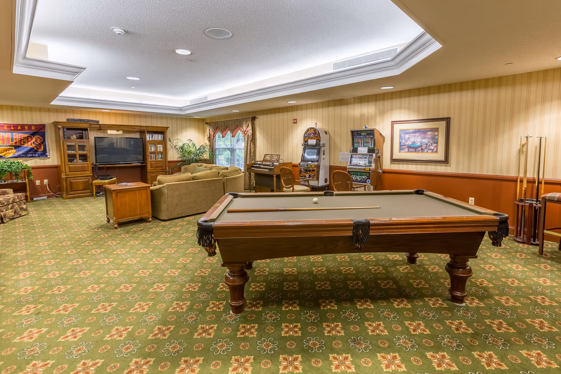 A recreational room with a pool table in the foreground, a seating area with couches and a large TV in a wooden entertainment center, two slot machines, a piano, and a Cleveland Cavaliers banner on the wall. The room has patterned green carpet, beige and brown walls, and a window with curtains.