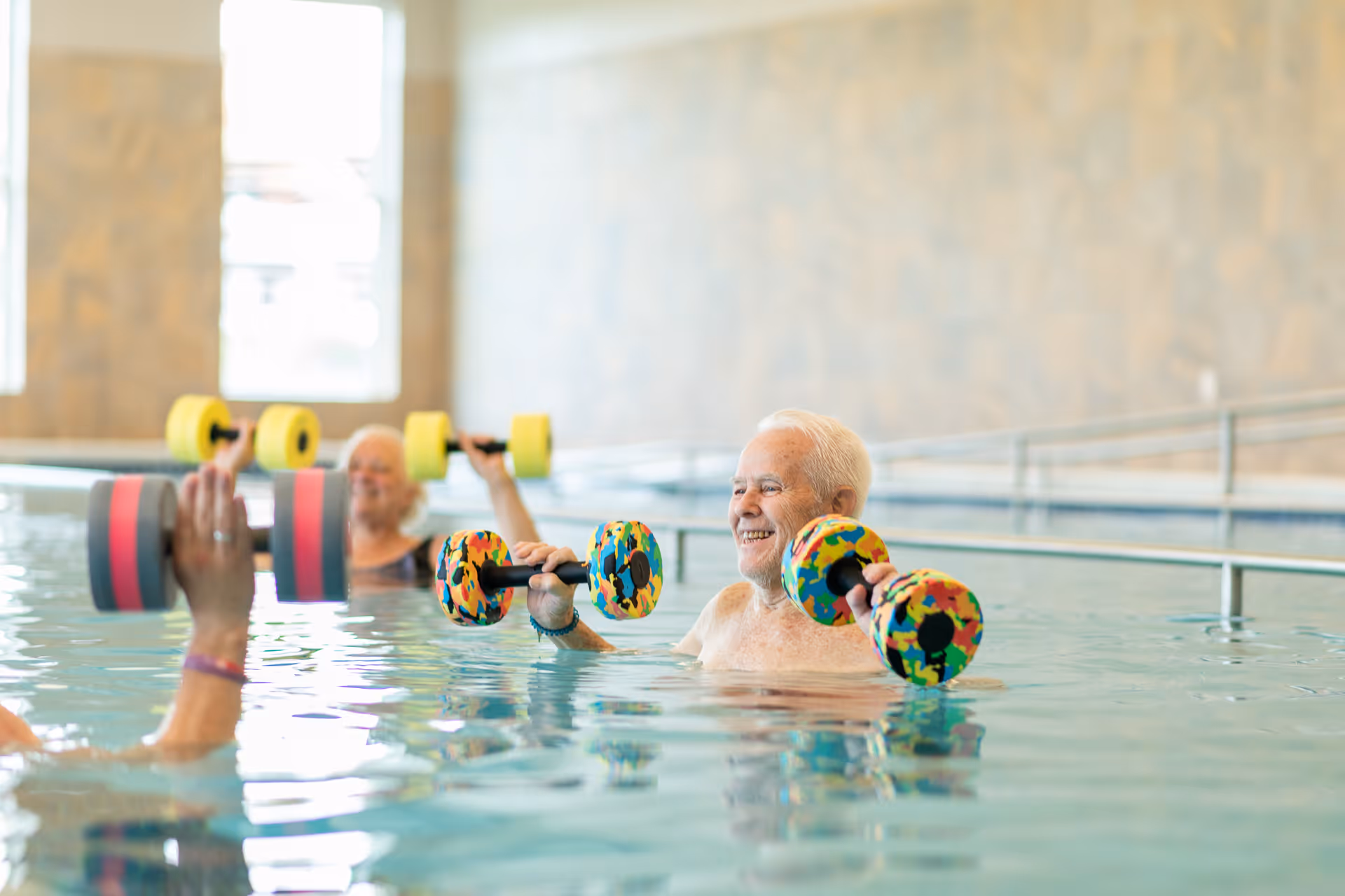 Three elderly individuals participating in a water exercise class in an indoor pool, each holding colorful foam dumbbells and smiling.