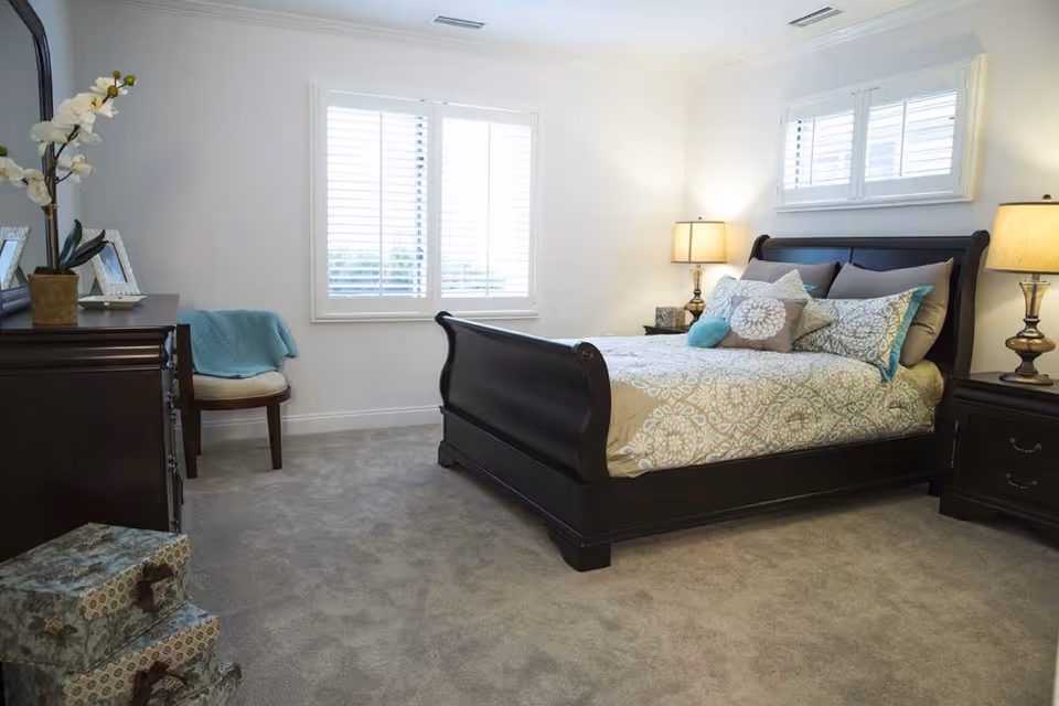 A bright bedroom with a dark wooden sleigh bed adorned with patterned bedding and multiple pillows. There are two matching bedside tables with lamps on either side of the bed. A window with white shutters is above the bed, and another window with shutters is on the adjacent wall. A wooden dresser with decorative items and a chair with a blue throw blanket are also visible. The room has light-colored walls and carpeted flooring.