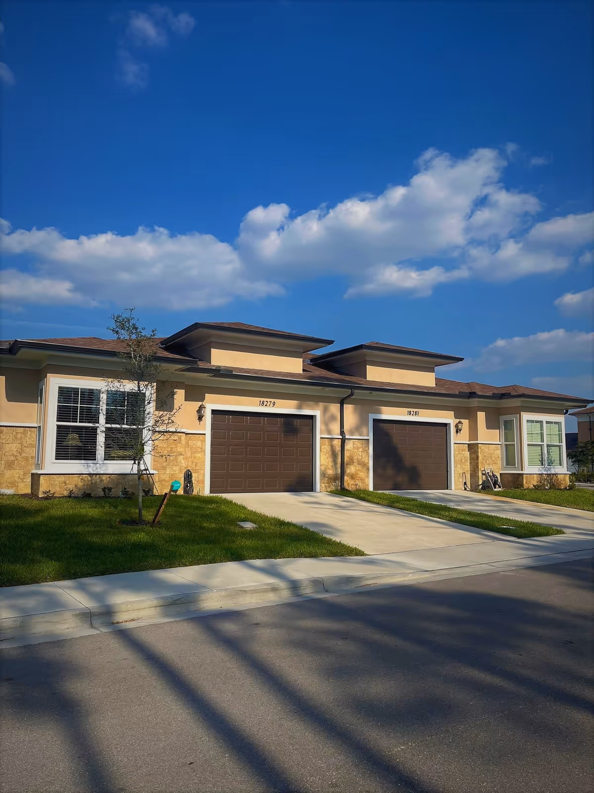 Exterior view of a single-story residential building with two attached garages, each with a brown garage door and a driveway. The building has beige and stone facade under a clear blue sky with some clouds. There is a small tree and green lawn in front of the building.