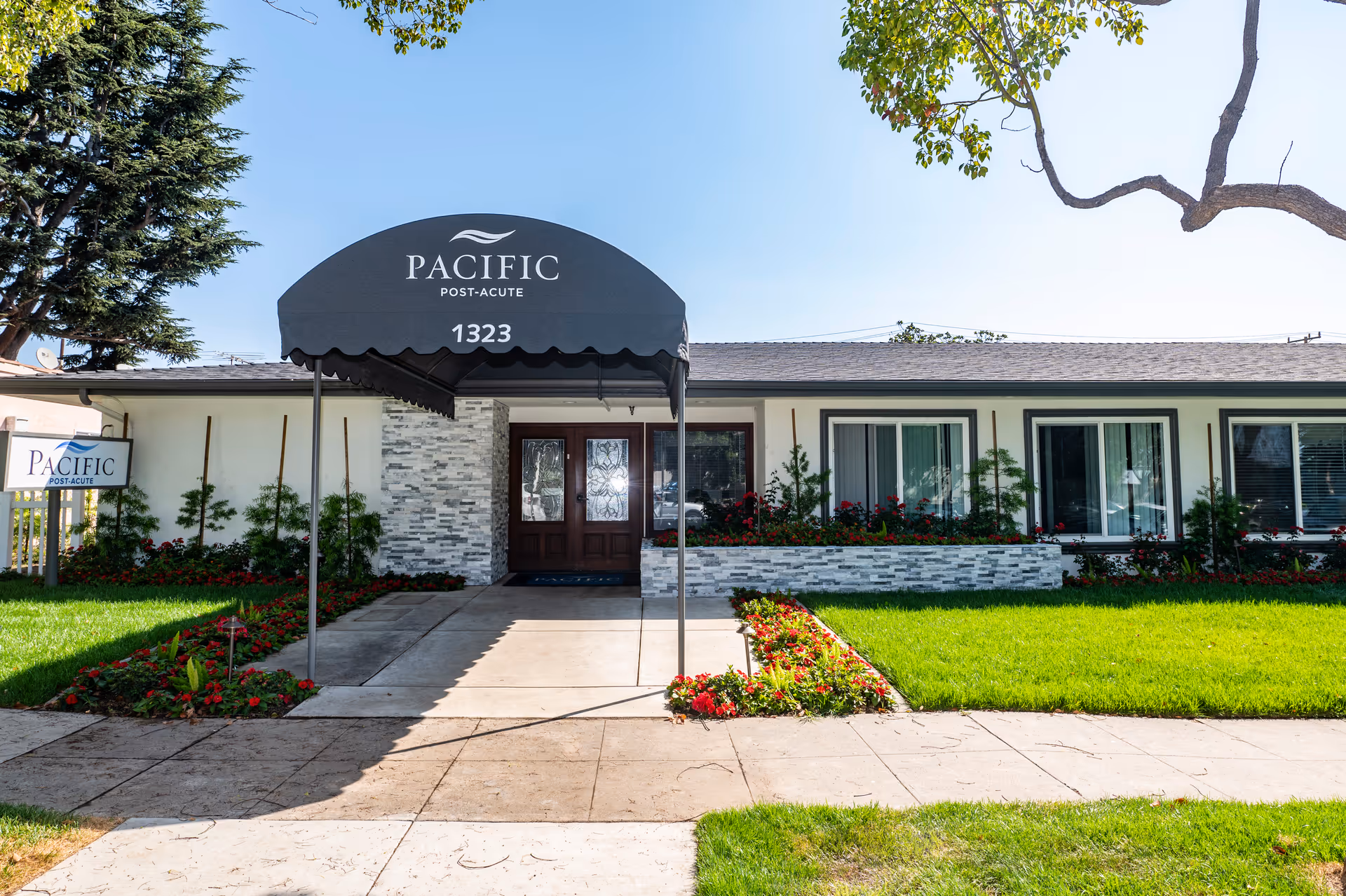 Front exterior and entrance of the Pacific Post-Acute building with a black awning reading 'PACIFIC POST-ACUTE 1323', landscaped lawn and windows.
