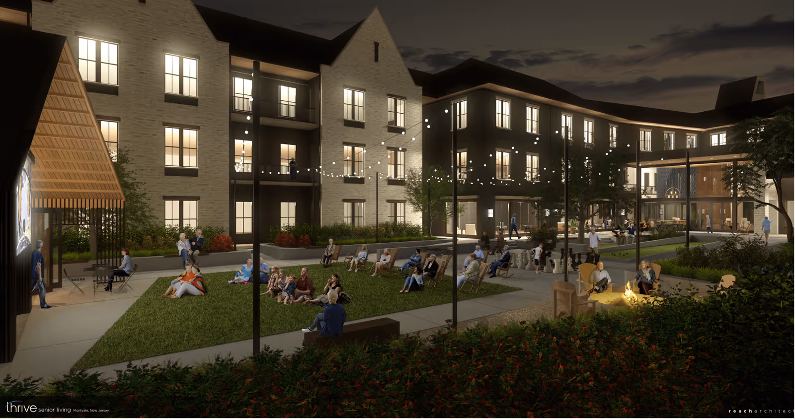 Nighttime view of an outdoor courtyard at a senior living facility with people sitting on grass and chairs watching a movie on a large screen. The courtyard is surrounded by a multi-story building with lit windows, string lights hanging above, and a fire pit area where two people are seated. There are trees, bushes, and pathways throughout the space.