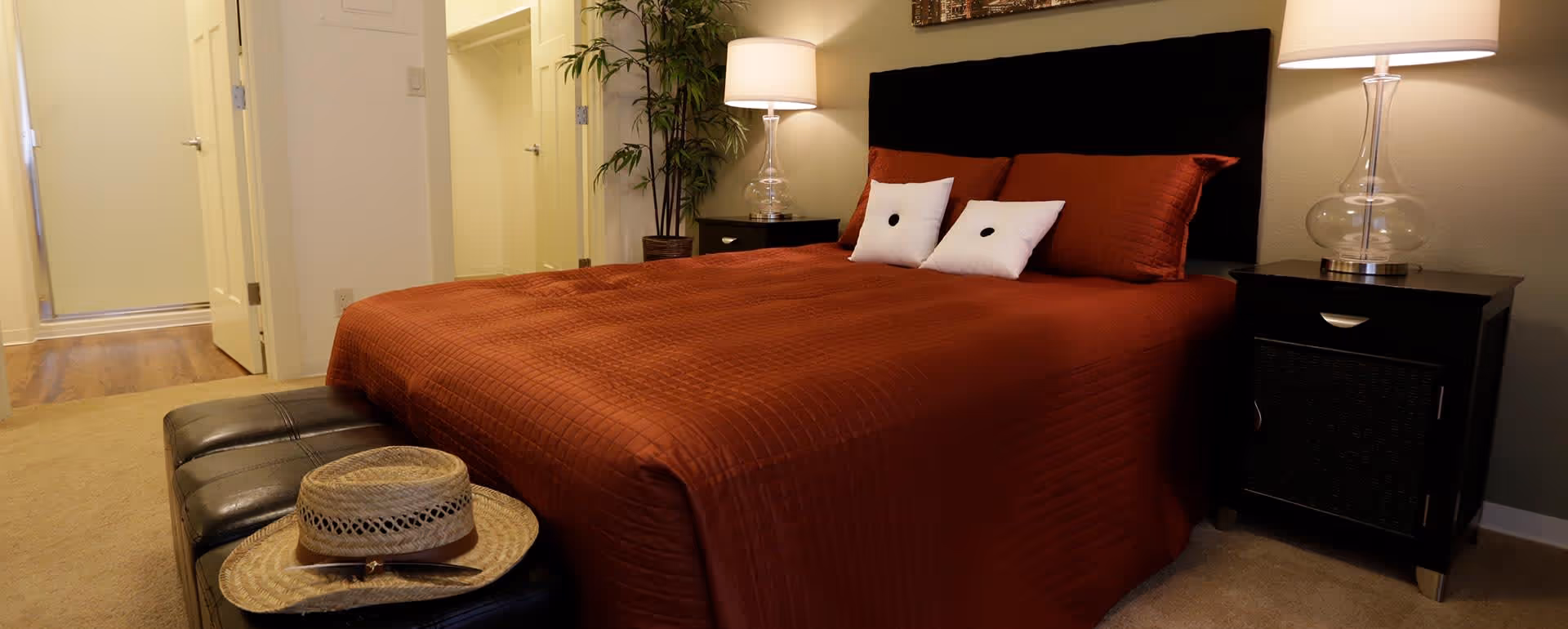 A bedroom with a bed covered in a rust-colored quilt and two white pillows with black buttons. There are two black nightstands on either side of the bed, each with a lamp. A black cushioned bench at the foot of the bed holds a straw hat. The room has beige carpet and light-colored walls, with a plant in the corner and an open doorway leading to another room.