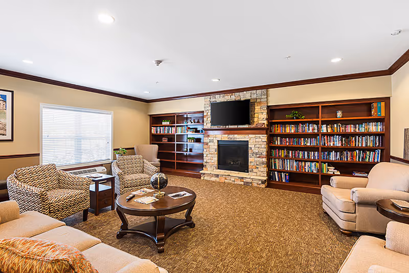 A cozy living room with beige walls and carpeted floor featuring a stone fireplace with a mounted flat-screen TV above it. On either side of the fireplace are wooden bookshelves filled with books and decorative items. The room has multiple comfortable armchairs and sofas arranged around a wooden coffee table with a globe and magazines on it. A large window with blinds allows natural light into the room.