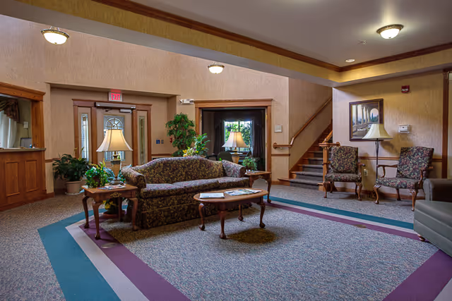 A cozy senior living facility common area with floral upholstered sofa and chairs, wooden coffee and side tables with lamps, potted plants, carpeted floor with a patterned border, a staircase leading upstairs, and framed artwork on the wall.