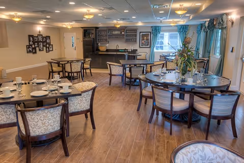 A dining room with multiple round and square tables set with plates, cups, and silverware. The room has wooden flooring, upholstered chairs, and a counter area with shelves and a refrigerator in the background. Large windows with blue curtains allow natural light to enter the space.