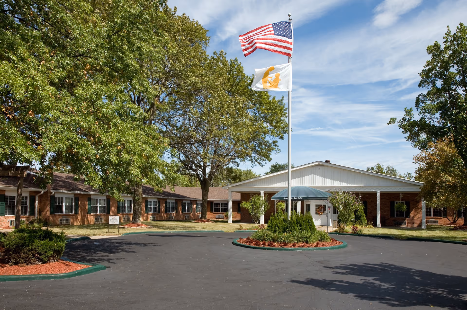 Single-story brick care center entrance with flagpoles, a circular driveway, and surrounding trees.