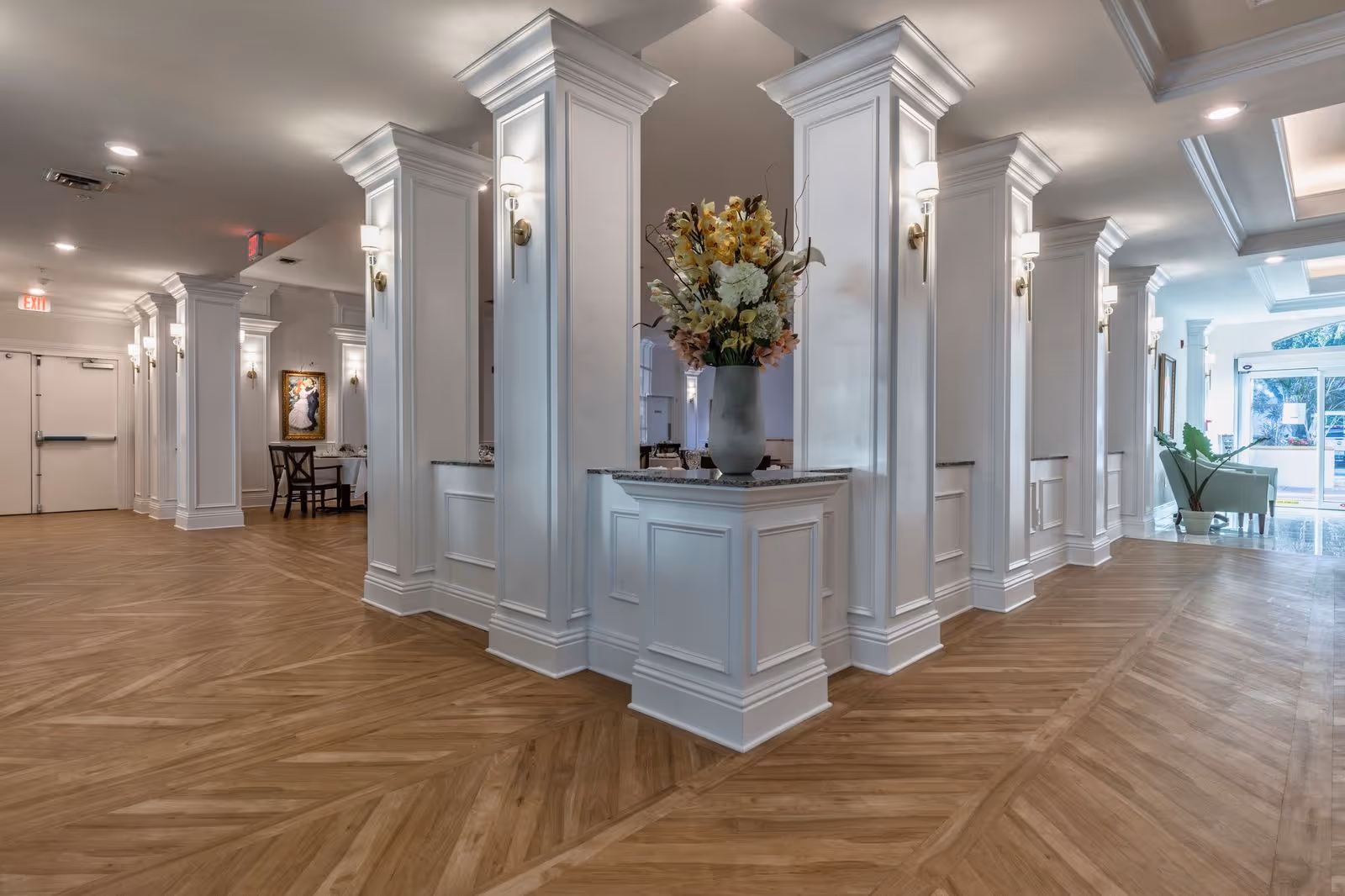 Elegant lobby interior with white columns, chevron wood floors and a floral arrangement on a central pedestal.